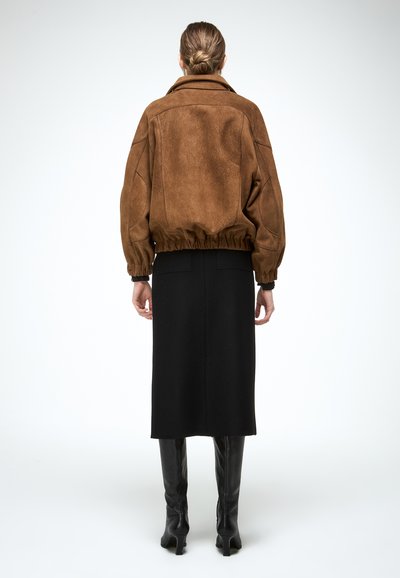 Žena s blond vlasmi zviazanými do drdolu, oblečená v hnedom semišovom saku, čiernej midi sukni a čiernych vysokých členkových čižmách, stojaca pred bielym pozadím.