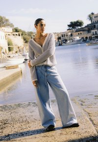 Csíkos kardigánt és bőszárú farmert viselő nő áll egy nyugodt kikötő mellett, ahol hajók és épületek láthatók lágy napsütésben.