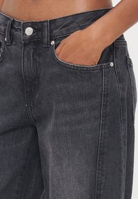 Jeans de ganga pretos com um corte relaxado, apresentando um fecho de botão, passantes para cinto e bolsos frontais. O tecido exibe uma textura e sombreamento sutis.
