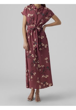 Femme portant une robe maxi fleurie bordeaux avec une ceinture nouée à la taille et des manches courtes, associée à des talons à brides transparentes, se tenant devant un fond gris.