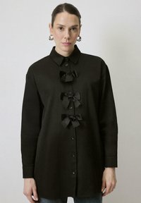 Zwarte blouse met drie strikaccenten aan de voorkant. Voorzien van een klassieke kraag en lange mouwen. Gladde stoftextuur.