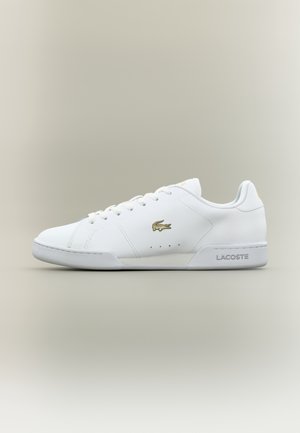 Biały niski sneaker Lacoste z złotym logo krokodyla, sznurowanym fasonem i oznaczeniem na podeszwie, pokazany z boku na neutralnym tle.