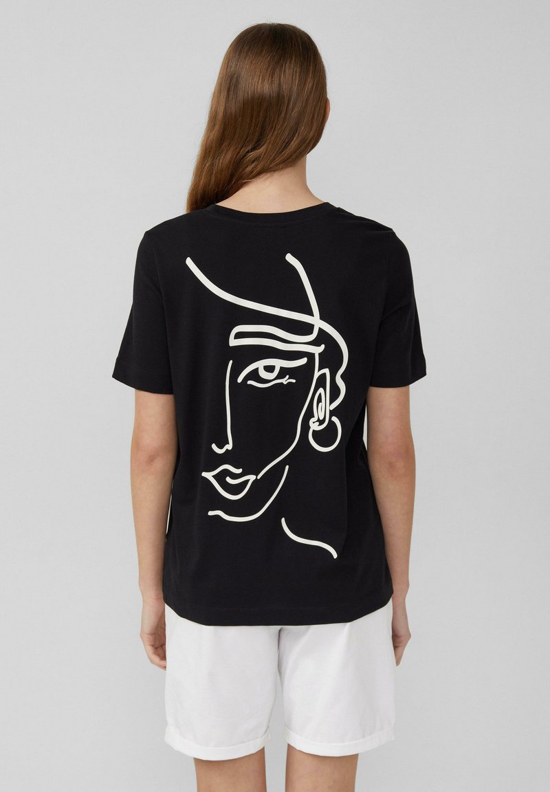 T-shirt in cotone nero con una linea bianca che rappresenta un volto sul retro. Il design include un orecchino come dettaglio. Maniche corte e colletto girocollo.