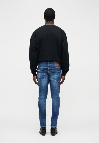 Άντρας που στέκεται με την πλάτη, φορώντας μαύρο φούτερ, μπλε τζιν με φθορές και πίσω τσέπες, και μαύρα μποτάκια αστραγάλου σε απλό λευκό φόντο.