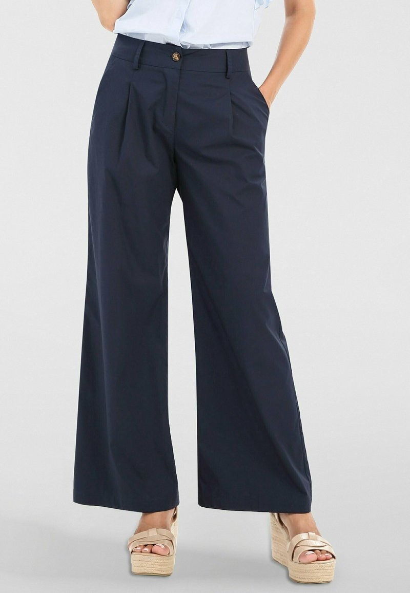 Femme portant un pantalon large bleu marine à plis sur le devant et des sandales beige à plateforme, debout avec une main dans la poche.