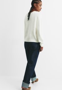 Weißer gerippter Strickpullover, dunkelblaue Jeans mit umgeschlagenem Saum und braunes Schuhwerk. Von hinten betrachtet, um Textur und Passform zu zeigen.