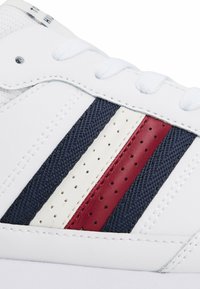 Witte sneaker met marineblauwe, rode en crèmekleurige gestreepte accenten. Heeft een geperforeerd ontwerp aan de zijkanten en is gemaakt van stevig materiaal met een textuurafwerking.