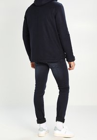 Felpa con cappuccio blu navy e cordino, abbinata a jeans scuri e sneakers bianche con dettagli azzurri. Vista posteriore.