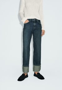 Jeans dritti blu scuro con risvolto, abbinati a un maglione lavorato a maglia color crema e a scarpe slip-on nere.