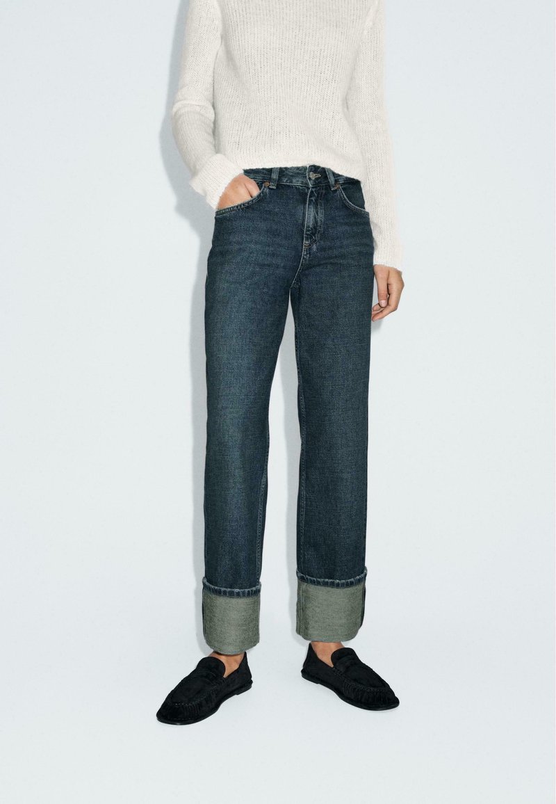 Jeans dritti blu scuro con risvolto, abbinati a un maglione lavorato a maglia color crema e a scarpe slip-on nere.