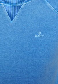 Jersey azul claro con cuello redondo de canalé azul. Presenta un pequeño logo blanco con "GANT" en el lado inferior izquierdo. Textura suave.