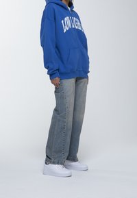 Felpa blu con zip e testo bianco, jeans chiari blu in vestibilità comoda e sneaker bianche. Presenta una tasca frontale e il cappuccio.