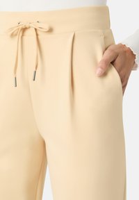 Béžové kalhoty s hladkou texturou, se stahovací šňůrkou v pase a dvěma bočními kapsami. Přehyby dodávají designu rozměr.