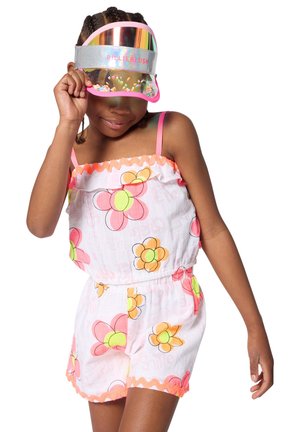 Jeune fille portant une combishort à motifs floraux et une visière réfléchissante ornée de paillettes, tenant la visière du bout des doigts et regardant vers le bas avec un léger sourire.