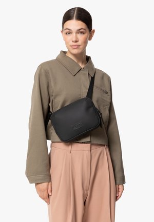 Sac à épaule noir zippé avec une finition mate lisse et un logo embossé, porté sur une veste collier verte et un pantalon rose clair.