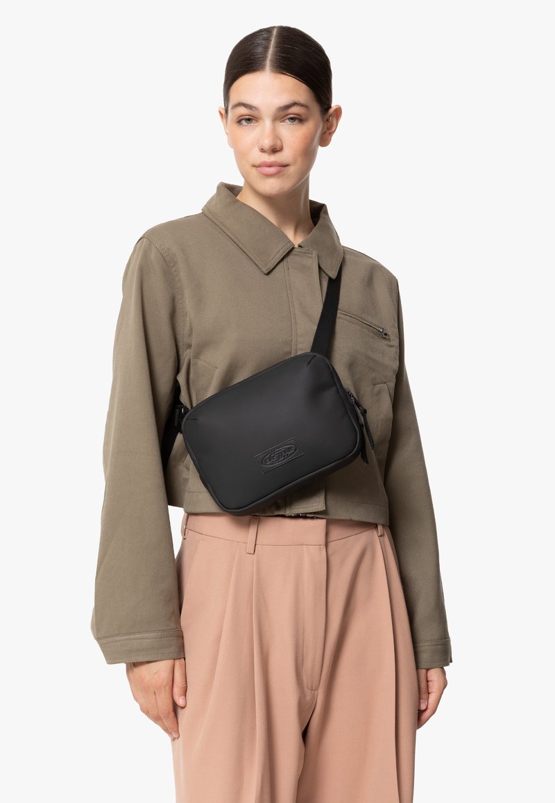Zwarte met rits afgesloten schoudertas met een gladde matte afwerking en een geëmbosseerd logo, gedragen over een groene colbert en lichtroze broek.