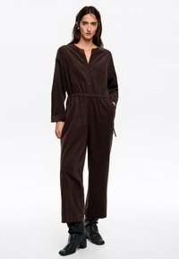 Mono de tela marrón con cuello alto, mangas largas, cintura elástica y bolsillos laterales. Se lleva con botas de tacón negras. Diseño simple y texturizado.