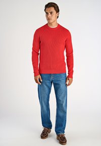 Uomo che indossa un maglione rosso a costine, jeans blu e scarpe in pelle marroni, in piedi contro uno sfondo chiaro e uniforme.