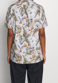 Camicia a maniche corte realizzata in tessuto leggero, con una stampa floreale tropicale e di uccelli in colori pastello su uno sfondo grigio chiaro.