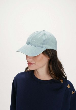 Femme portant une casquette de baseball bleu clair avec le texte « PEACE & MILA » et un haut bleu marine avec des boutons dorés sur l'épaule, sur un fond uni.
