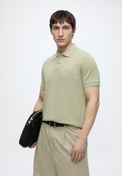 Homme portant un polo vert clair et un pantalon beige tient une pochette noire zippée sur un fond blanc uni.