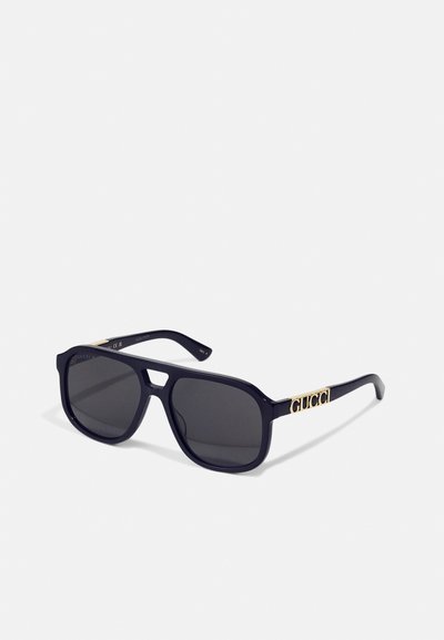 Lunettes de soleil en plastique noir avec des verres foncés, de forme carrée, arborant le logo doré "Gucci" sur la branche. Fini mat avec un design élégant.