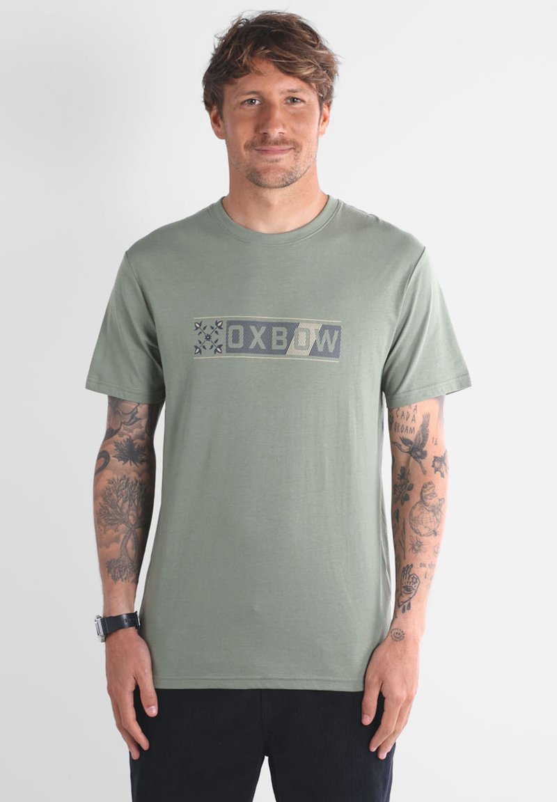 T-shirt en coton vert olive avec un logo rectangulaire "OXBOW" en bleu foncé et gris clair, à manches courtes et col rond.