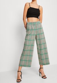Femme portant un pantalon large à carreaux taille haute avec des accents verts, un top noir à bretelles spaghetti et des sandales à talons hauts noires.