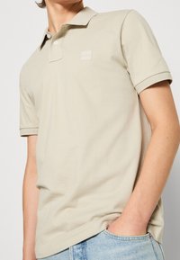 Polo beige en coton avec un col, des manches courtes et un logo tonal carré sur la poitrine gauche. Texture douce et coupe décontractée.