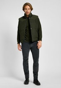 Gilet imbottito verde con colletto in velluto a coste, abbinato a una camicia a maniche lunghe verde scuro e pantaloni grigio scuro, indossato con scarpe nere.