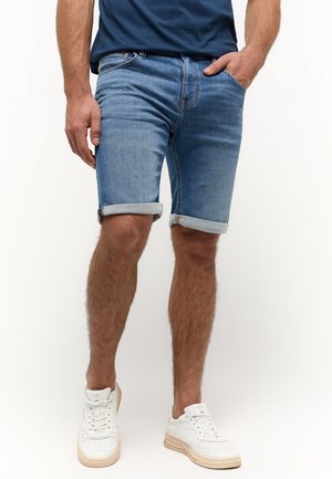 Homme portant un short en jean bleu retroussé, des baskets blanches et une chemise bleu foncé, une main dans la poche.