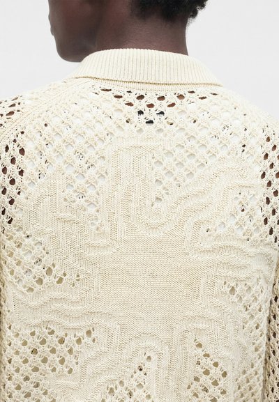 Άτομο που φοράει κρεμ πουλόβερ με πλεκτό σχέδιο ηλιακών ακτίνων και διάτρητες λεπτομέρειες στην πλάτη, γυρισμένο από την πλάτη.