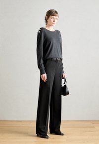 Pull en maille gris avec des boutons d'épaule, large pantalon noir, et un petit sac à main noir. Tissu lisse avec une coupe détendue. Chaussures noires à bout carré.