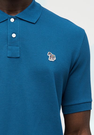 Μπλε πόλο μπλούζα από υφασμάτινη υφή, με γιακά και δύο κουμπιά. Μικρό πολύχρωμο λογότυπο ζέβρας είναι κεντημένο στο στήθος.