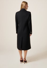Cappotto lungo in misto lana nero con design sartoriale, schiena spaccata e dettagli con bottoni dorati sulle maniche. Silhouette elegante e strutturata.