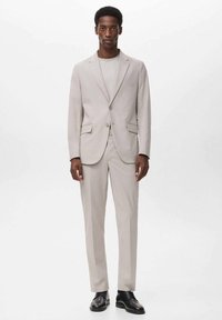 Light beige suit with structured blazer and flat-front trousers, featuring two front pockets and button closure. Simple crew neck shirt underneath.