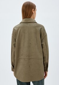 Camisa chaqueta de ante sintético verde oliva con pliegue en la espalda, puños abotonados y cuello clásico, que presenta una textura suave.