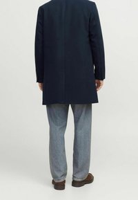 Marinblå rock med rak skärning, två knappar och sidventiler, kombinerad med ljusblå denimbyxor och bruna skor, sedd bakifrån.