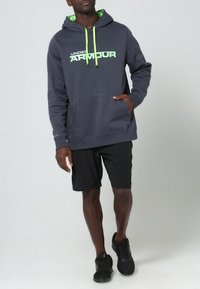 Sudadera con capucha gris con detalles y logotipo en verde, con un bolsillo frontal, combinada con pantalones cortos negros y zapatillas deportivas negras.