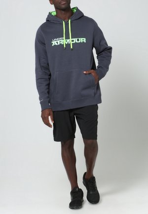 Grauer Kapuzenpulli mit grünen Akzenten und Logo, ausgestattet mit einer Fronttasche, kombiniert mit schwarzen Shorts und schwarzen Sportschuhen.