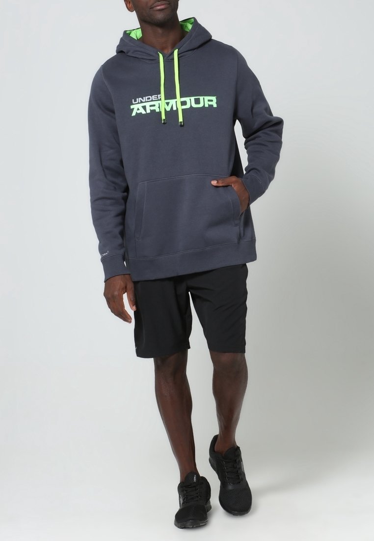 Sudadera con capucha gris con detalles y logotipo en verde, con un bolsillo frontal, combinada con pantalones cortos negros y zapatillas deportivas negras.