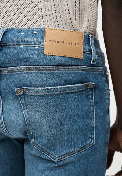 Κοντινή λήψη της πίσω τσέπης και της ζώνης μιας μπλε τζιν με δερμάτινη ταμπέλα σε ταμπά χρώμα που γράφει «Tiger of Sweden», φορεμένη με ανοιχτό μπεζ πλεκτό τοπ.