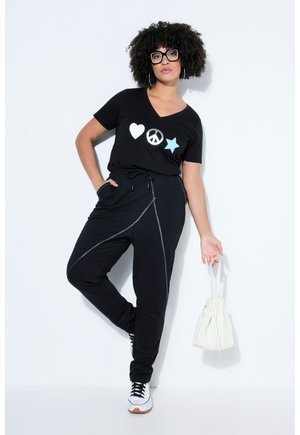 Vrouw met krullend haar, gekleed in een zwart T-shirt met hart-, vrede- en stericonen, zwarte broek, witte sneakers, en een kleine witte handtas vasthoudend.