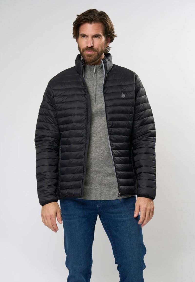 Schwarze Steppjacke mit horizontalem Steppmuster, hohem Kragen und Reißverschluss, getragen über einem hellgrauen Pullover und blauen Jeans.
