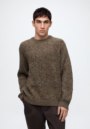 Jeune homme aux cheveux courts et foncés, portant un pull en tricot marron et un pantalon foncé, debout avec une main dans la poche devant un fond uni.