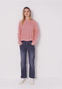Rosa Pullover mit langen Ärmeln in lockerer Passform, kombiniert mit dunkelblauen Jeans mit geradem Bein. Das Model trägt weiße Sneakers und präsentiert einen lässigen Look.