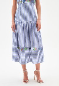 Sininen ja valkoinen raidallinen midi-mekko, jossa on kukkakirjontaa vyötärön ja helmasta, käytetty valkoisten korkokenkien kanssa valkoista taustaa vasten.