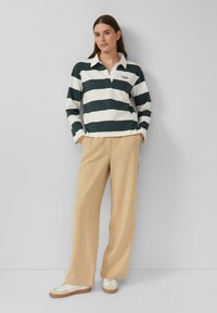 Vrouw staat tegen een grijze muur en draagt een zwart-wit gestreept shirt met lange mouwen, beige wijduitlopende broek en witte sneakers.