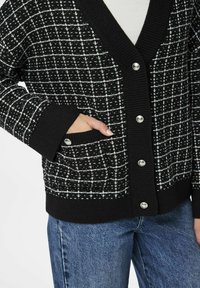 Mustrita mustvalge kardigan V-kaelusega, soonikkoes varrukatega ja taskuga. Omab metallnuppe ja pehmet, kudumist tekstuuri.