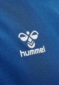 Mėlynas audinys su baltu "hummel" logotipu, kuriame stilizuoti sparnai ir geometrinė raštas, esantis po tekstu. Lygi tekstūra.